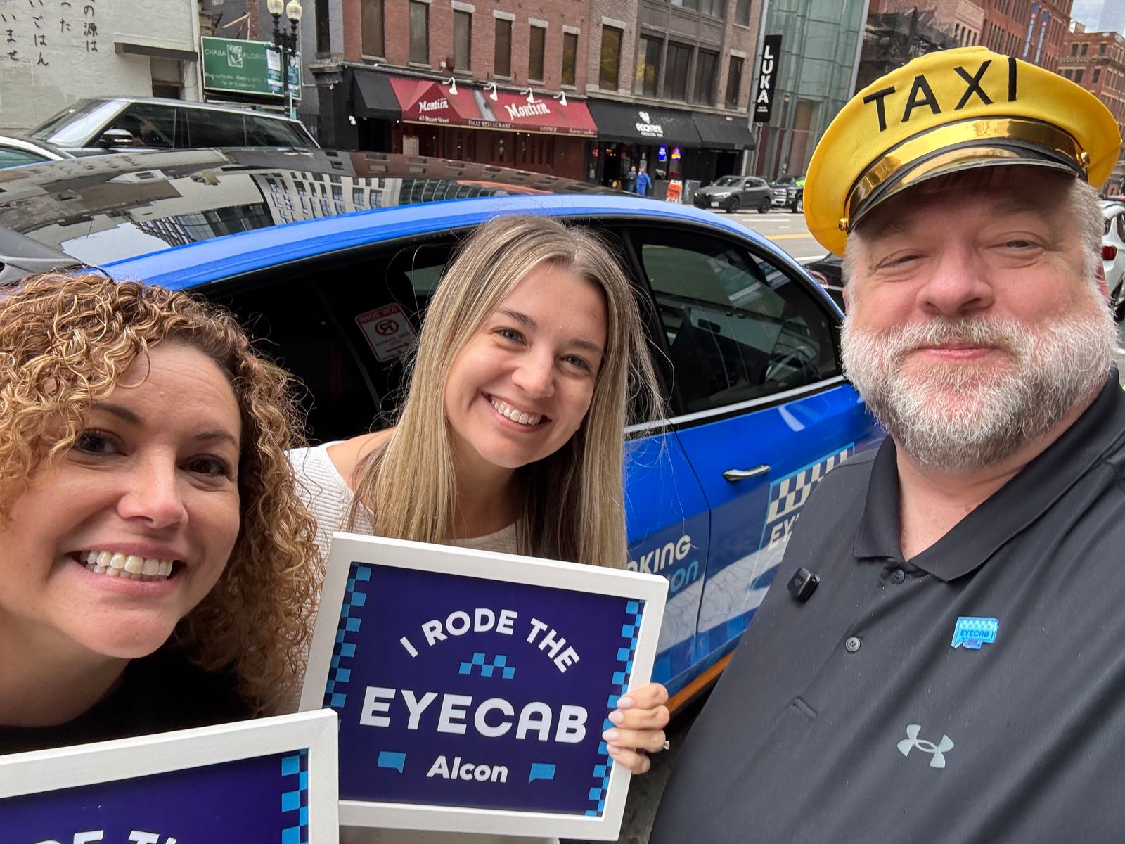 The EyeCab driver in a TAXI cap taking a selfie with two riders beside the cab